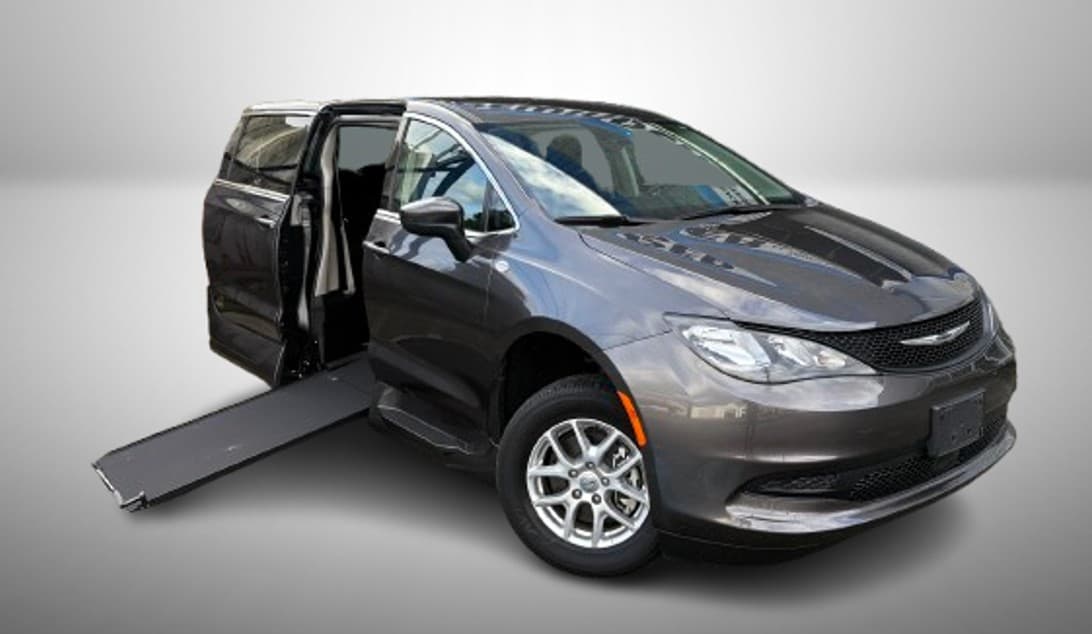 Granite grey Chrysler Grand Caravan with the VMI Northstar E side-entry conversion, front three-quarter view, sliding passenger door open and the in-floor ramp lowered to the ground